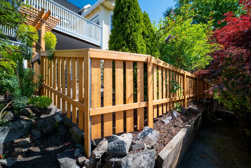 A wooden slat fence surrounds a garden with rocks and various green plants beside a house with a white balcony and trees in the background under a blue sky.