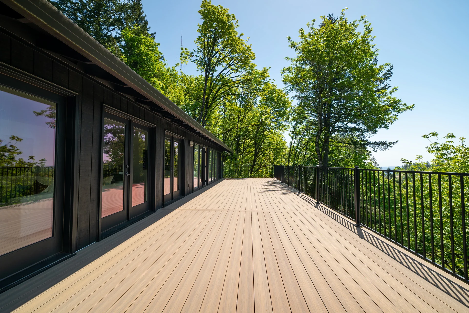 A spacious wooden deck with black metal railings extends along a modern dark-paneled building, surrounded by lush green trees under a clear blue sky.