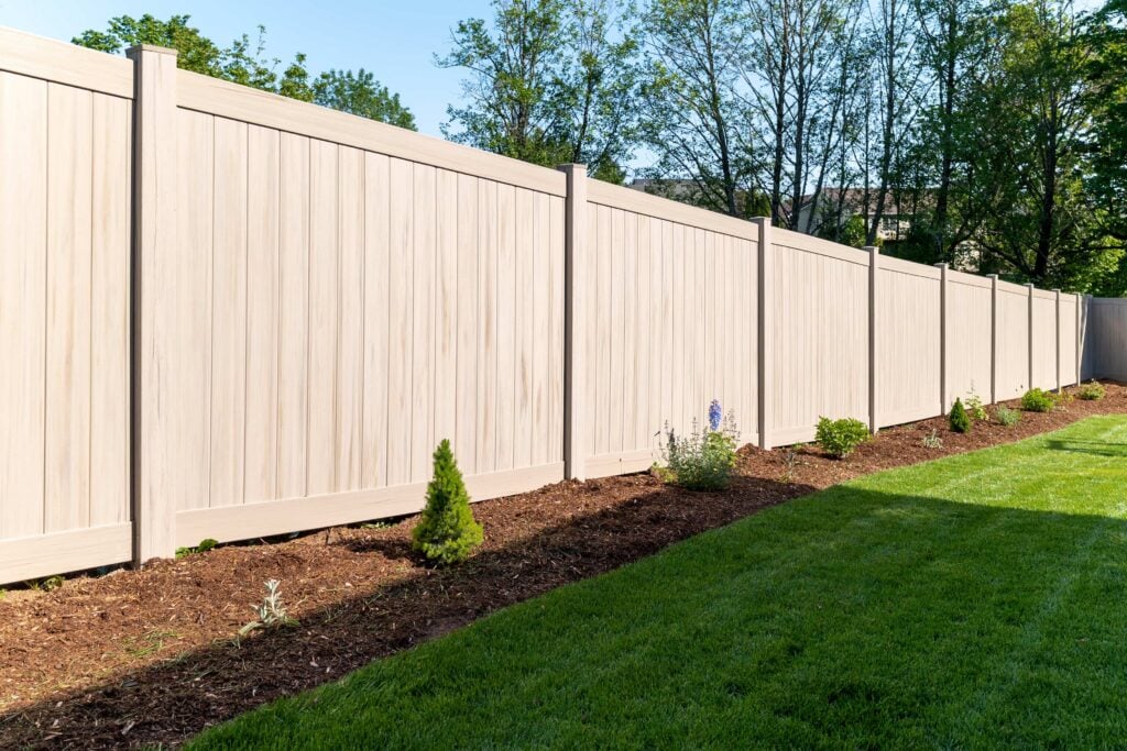 A tall, light-colored wooden privacy fence borders a neatly maintained backyard with green grass and young plants along a mulched garden bed. Trees are visible in the background.