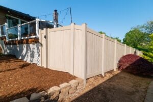 Tan privacy fence installed along the edge of a backyard, next to a raised deck and a garden bed with mulch.