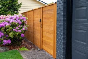 A horizontal wooden privacy fence with a gate connects to a dark grey brick house wall, next to a large blooming rhododendron bush.