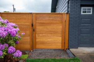 A horizontal cedar wood fence and gate next to a flowering purple rhododendron bush and a dark gray brick garage.