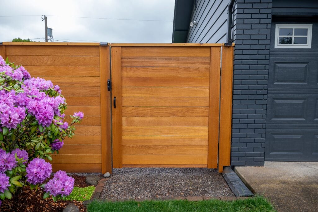 A wooden fence and gate stand next to a dark gray house and garage; blooming pink flowers and green grass are in the foreground.