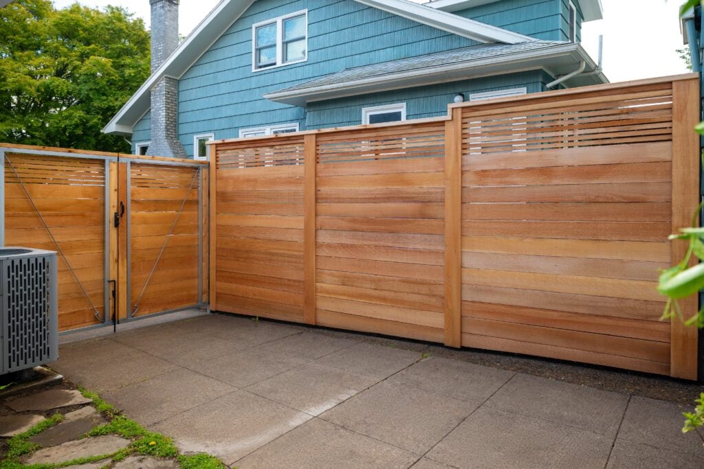 A modern wooden privacy fence with a metal gate encloses a small paved patio area beside a blue house. Lush green trees and an air conditioning unit are visible in the background.