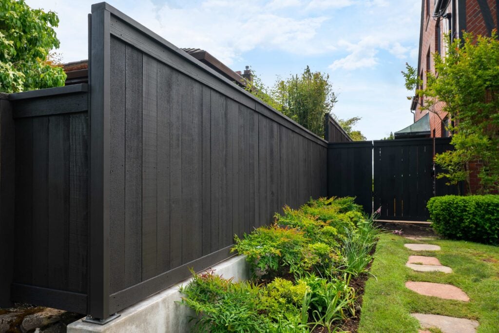 A modern black wooden fence lines a backyard, with green plants and a stone path running beside it. A brick house and trees are visible in the background under a partly cloudy sky.