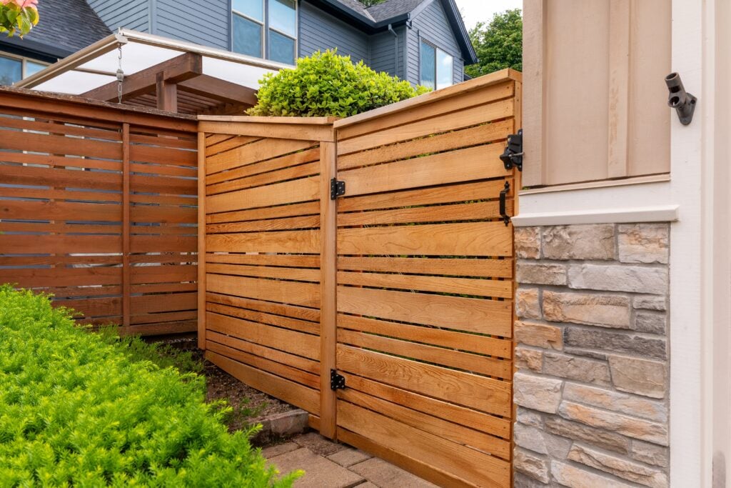 A wooden fence and gate with horizontal slats encloses a yard next to a house with stone and beige siding. Green bushes and another building are visible in the background.