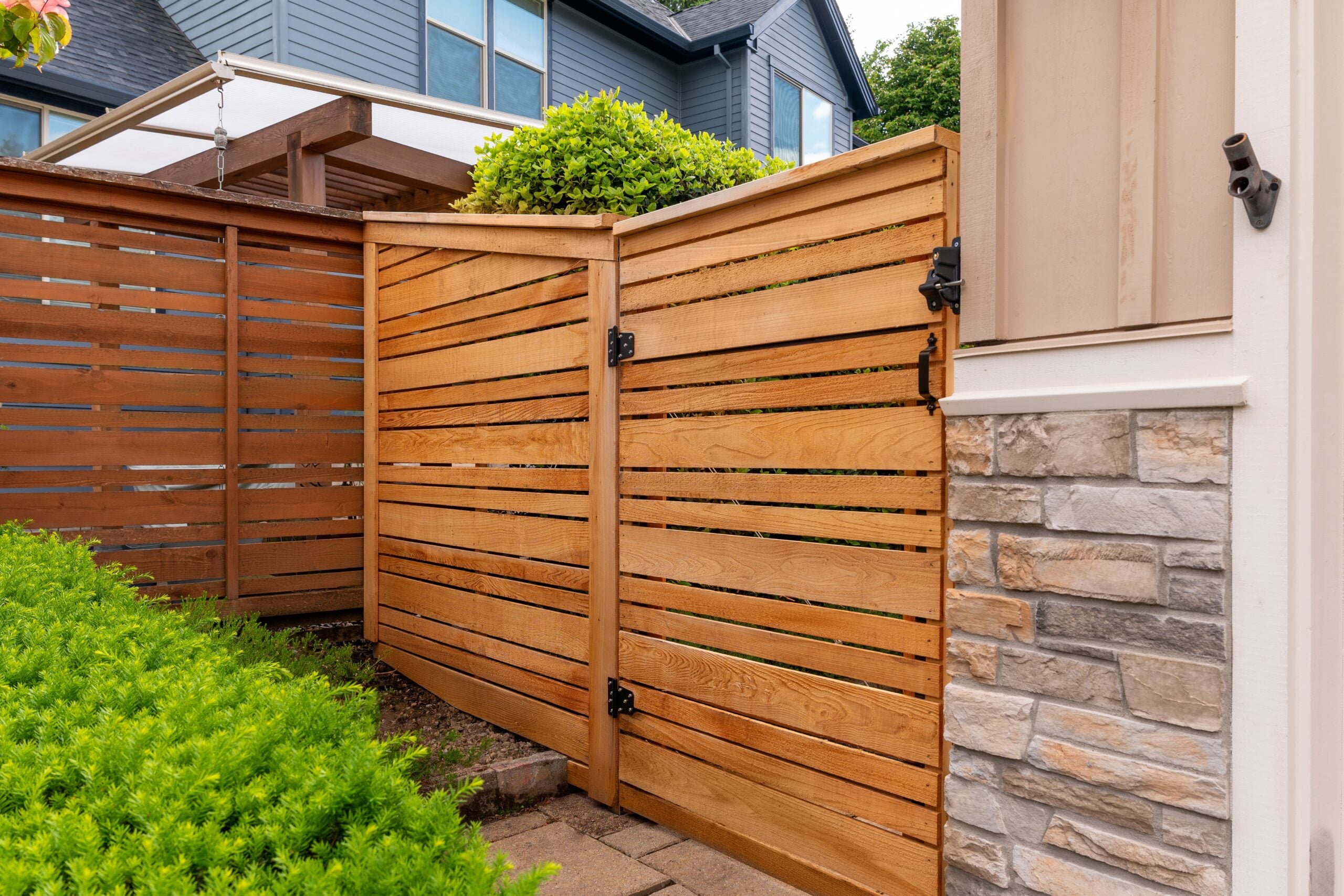 A wooden fence and gate with horizontal slats encloses a yard next to a house with stone and beige siding. Green bushes and another building are visible in the background.