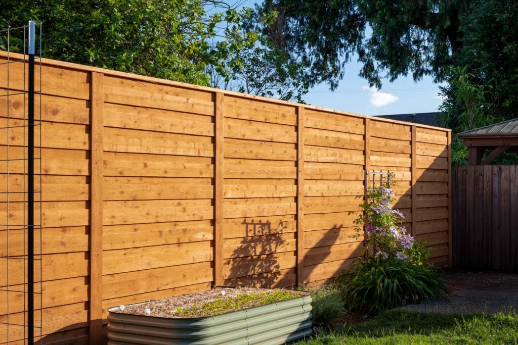 A tall wooden fence in a backyard with sunlight casting shadows, a raised garden bed in front, and some flowering plants and greenery along the fence.