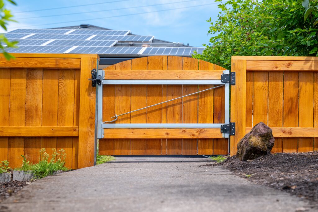 A wooden gate with metal hinges is partially open across a pathway. There is a large rock beside the gate, and solar panels are visible on the roof of a house in the background. Green plants line the path.