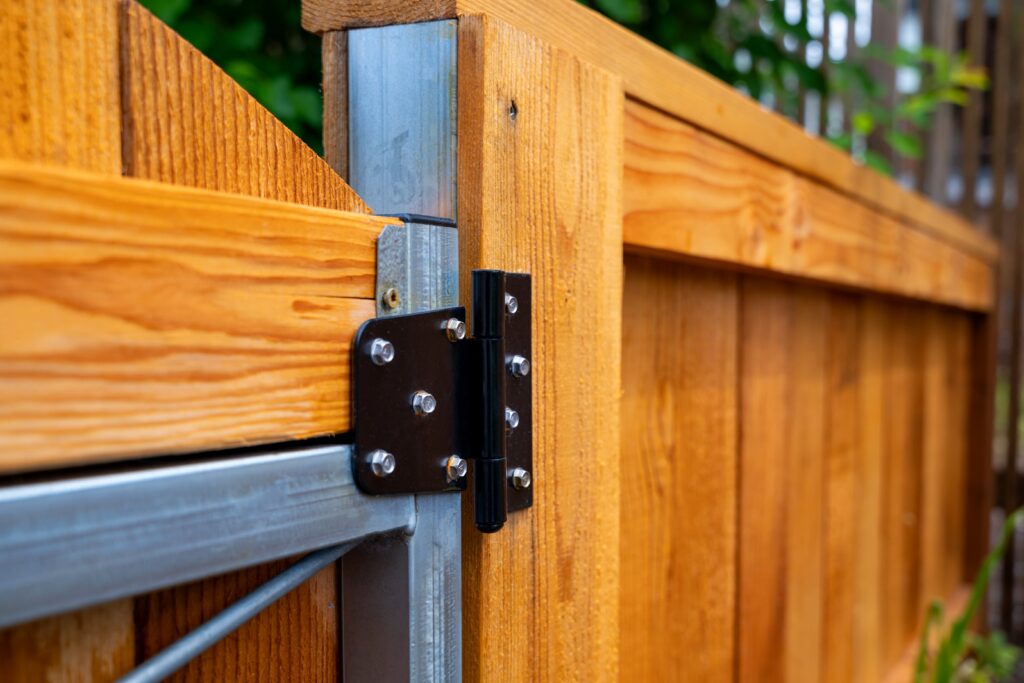Close-up of a metal hinge connecting a wooden gate to a wooden fence, showing the screws and metal frame, with greenery and fence detail in the background.