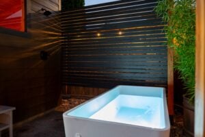 An illuminated outdoor cold plunge tub sits on a patio next to a horizontal slat privacy screen and a tall bamboo plant.