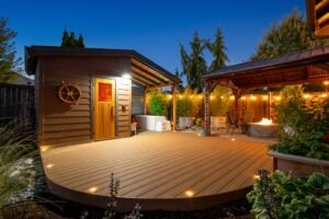 A curved wooden deck featuring a sauna with a ships wheel on the exterior, a soaking tub, and a covered patio area with a fire pit at dusk.