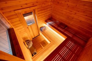 A high-angle view of a cedar sauna interior with L-shaped benches, a glass-paneled door, and a heater filled with stones.