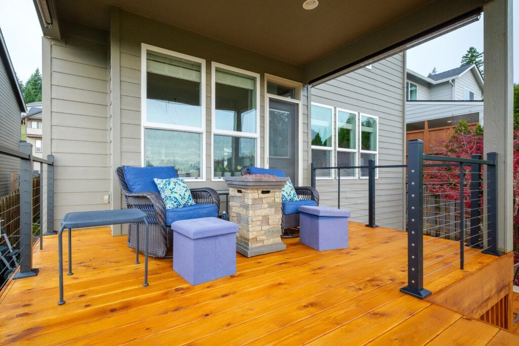 Modern backyard deck with wood flooring, wicker chair, blue cushions, and a stone fire pit. Glass-paneled railing surrounds the seating area. House and neighboring homes visible in the background.