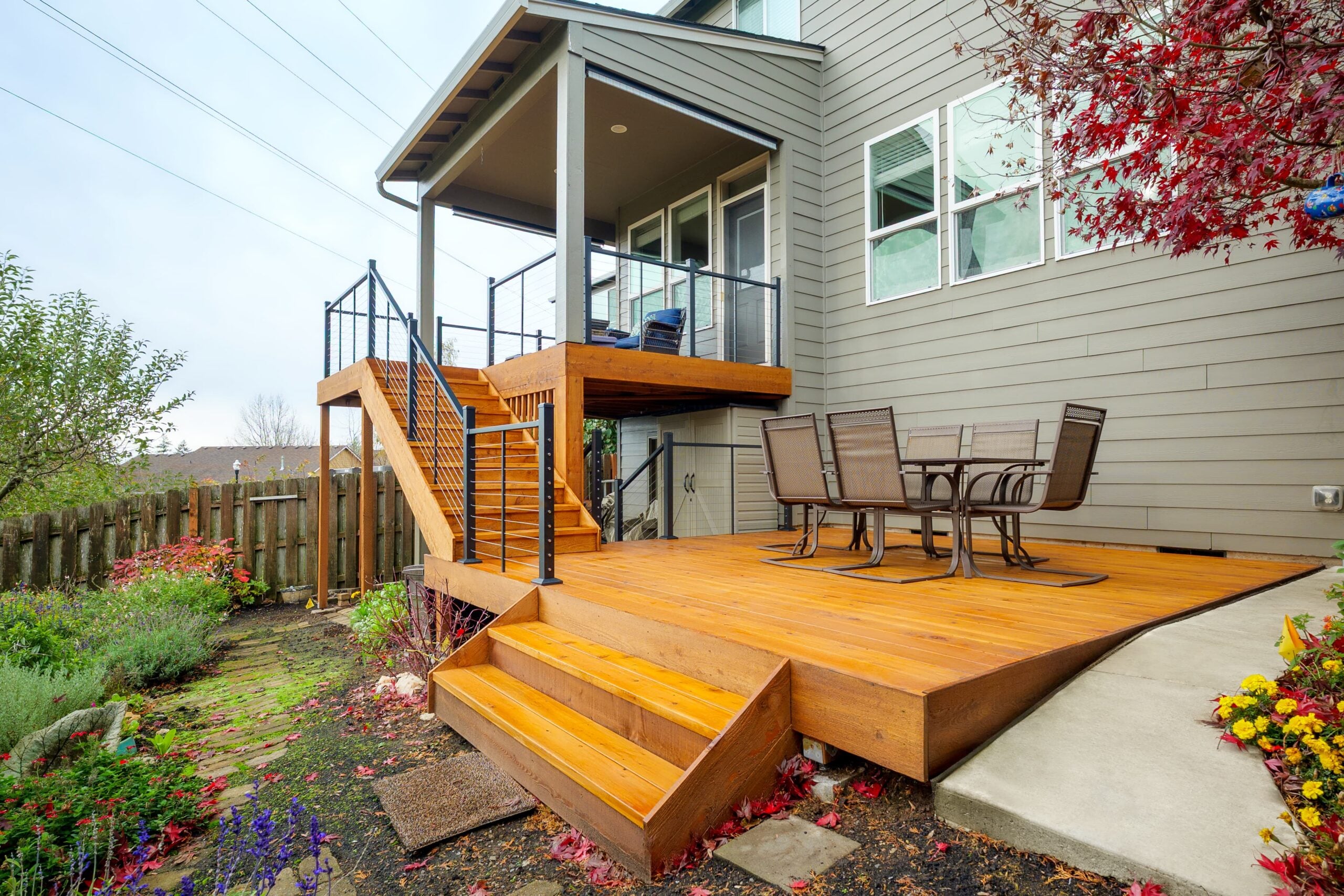 A wooden backyard deck with steps, a patio table and chairs, metal railing, garden beds, and red-leaved tree next to a gray house. An upper deck with seating overlooks the yard.