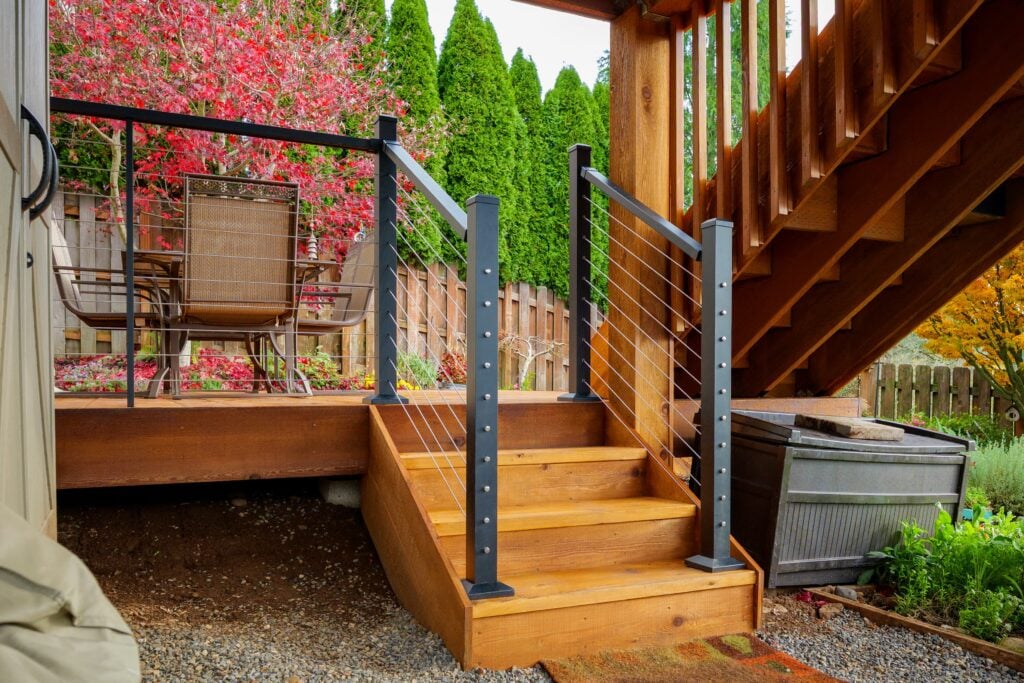 Wooden stairs with a black metal cable railing lead to a raised deck with an outdoor dining table and chairs. Lush greenery and red foliage are visible in the background.