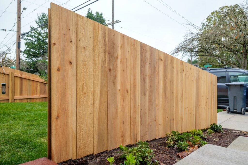 A newly installed wooden fence separates a grassy yard from a driveway with a parked car and trash bin. Small plants line the base of the fence, and power lines are visible in the background.