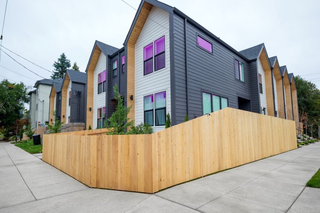 A row of modern townhouses with grey and white siding and purple-tinted windows, enclosed by a tall wooden fence.