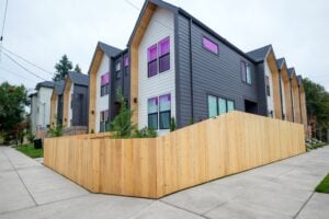 A row of modern townhouses with grey and white siding and purple-tinted windows, enclosed by a tall wooden fence.