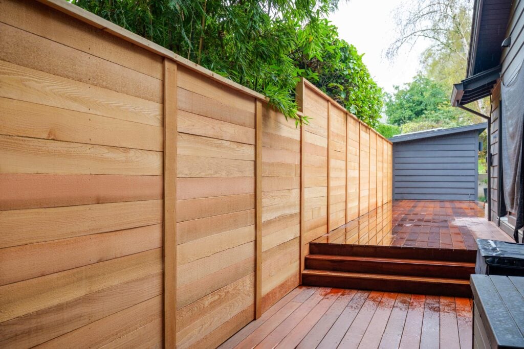 A wooden privacy fence lines a backyard deck with steps, leading to a gray shed. Green trees and plants overhang above the fence.