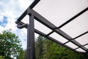 Black metal patio cover with a translucent roof and an integrated downspout, set against a background of trees and blue sky.