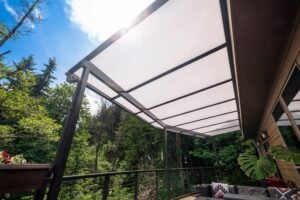 A patio covered by a translucent roof structure attached to the side of a house, overlooking a wooded area.