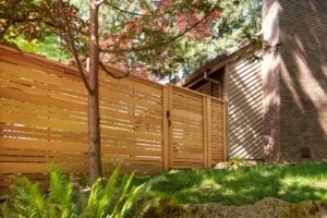 A horizontal wood slat fence and matching gate installed next to a house with brick siding and green foliage.