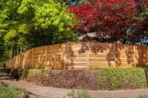 A horizontal wood slat fence sits atop a stone retaining wall in front of green and red trees.