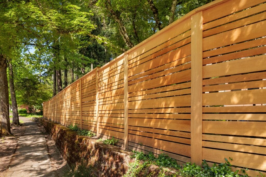 A tall wooden fence with horizontal slats runs alongside a shaded sidewalk, surrounded by green trees and plants on a sunny day.