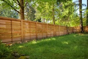 A horizontal wooden slat fence stands in a backyard bordered by green trees and grass.