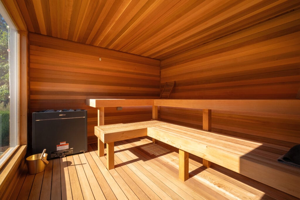 Wood-paneled sauna room with two-tiered benches, a black heating stove, a wooden bucket with a ladle, and sunlight streaming through a large window on the left side.