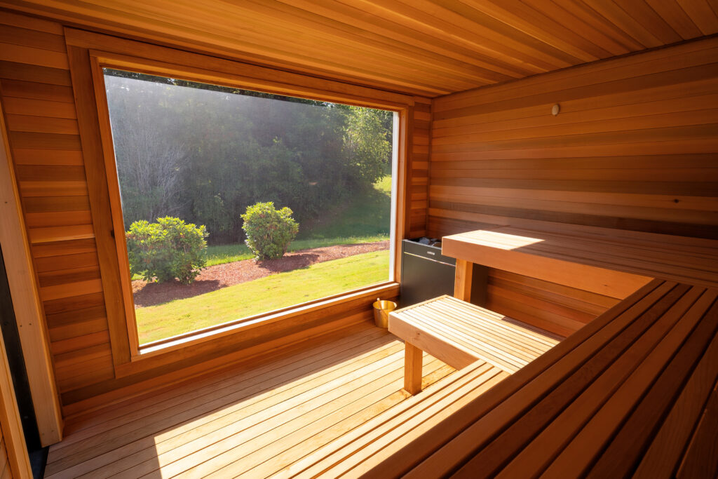 A wooden sauna with a large window offers a view of green grass, bushes, and trees outside, with sunlight streaming in and benches along the walls.