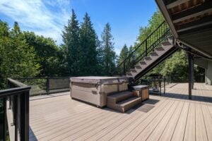 A hot tub with a cover sits on a wooden deck next to an outdoor staircase and a barbecue grill.