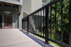 A wooden deck with a black metal railing overlooking a wooded area next to a house exterior.