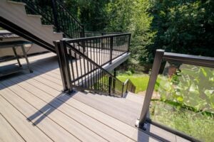 A multi-level outdoor deck featuring light-colored wood-look planks and black metal railings with glass panels, with stairs leading down to a green backyard.