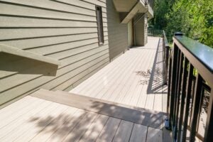A wooden deck with a staircase along the side of a house, featuring a dark metal railing overlooking a wooded area.