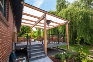A raised wooden deck with a translucent roof, featuring a patio heater, furniture, and short stairs leading to a backyard.