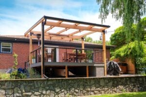 A backyard deck with a wooden pergola attached to a brick house, featuring outdoor dining furniture and a stone retaining wall.