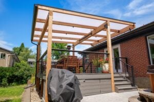 A wooden patio deck with a translucent roof, featuring a dining set, a covered grill, and steps leading to a brick house.