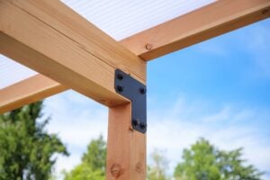 A black L-shaped metal bracket securing a corner connection between two wooden beams of a pergola.