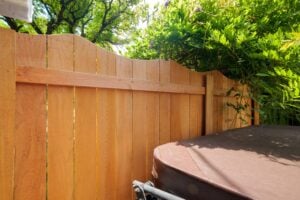 A wooden fence with a scalloped top sits beside a covered hot tub in a backyard with green foliage.