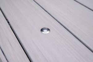 A small, circular, silver in-lite branded deck light recessed into gray wooden decking.