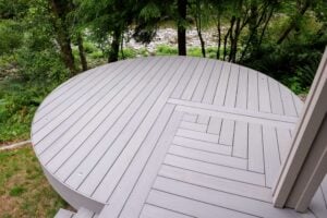 A circular grey deck with a geometric herringbone inlay, situated outdoors in a wooded area near a rocky stream.