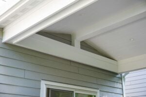 Close-up of a house exterior showing light gray horizontal siding, a white window frame, and a white covered porch ceiling with support beams.