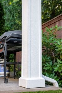 A white porch column on a stone patio, with a black outdoor griddle to the left and a downspout extending into a garden.