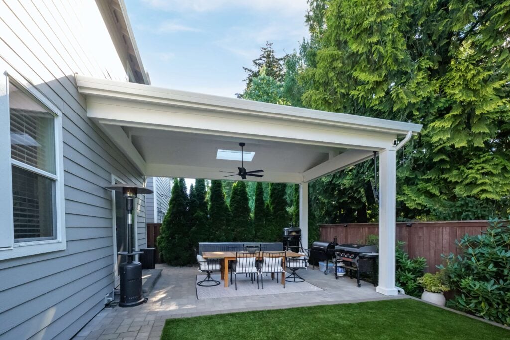 A covered backyard patio featuring a dining set with four chairs, a freestanding outdoor heater, and two grills.
