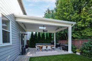 A covered backyard patio featuring a dining set with four chairs, a freestanding outdoor heater, and two grills.