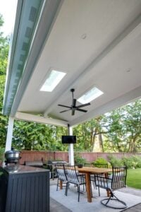 A covered outdoor patio featuring a dining table with chairs, a television mounted on a post, and a grill setup, sheltered by a roof with two skylights and a ceiling fan.