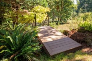 A composite deck walkway leading to a bridge in a garden setting surrounded by trees and ferns.