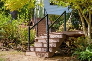Outdoor wooden deck stairs with black metal railings and cable balusters, nestled in a wooded backyard setting.